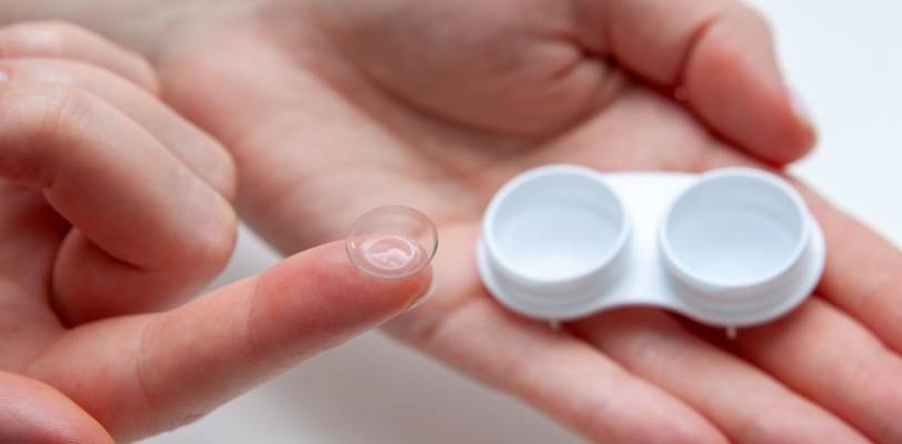 A single contact lens on the end of a finger, with a contact lens case on an open hand in the background.