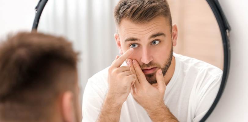 A man looking in the mirror to insert a contact lens in his eye.