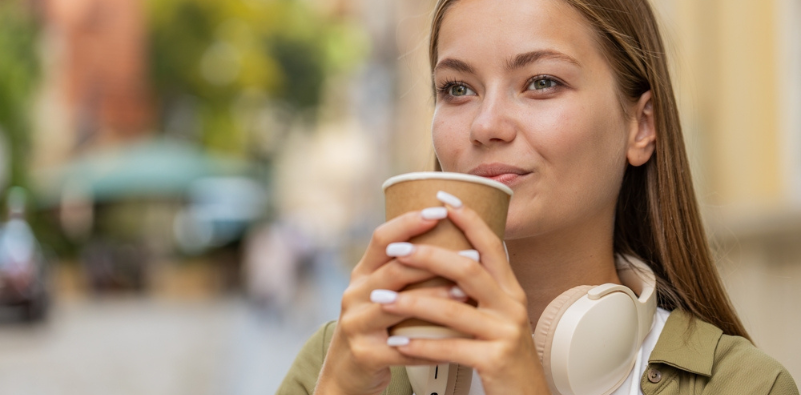 A young woman outside with headphones around her neck, holding a disposable brown coffee cup.