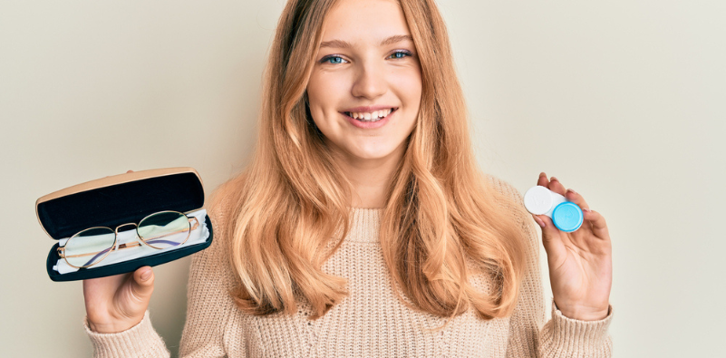 A young woman holding a pair of glasses in a case in one hand and a contact lens storage container in the other hand