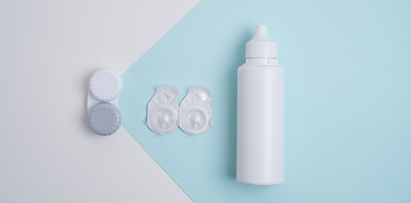 A white and light blue surface with a contact lens storage container on the left, a pair of contact lenses in the centre and a bottle of contact lens solution on the right