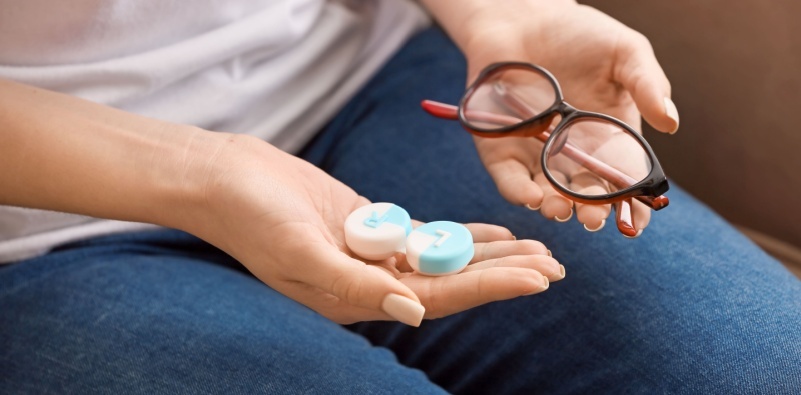A close up of a woman holding a pair of glasses in one hand and a contact lens storage pot in the other hand