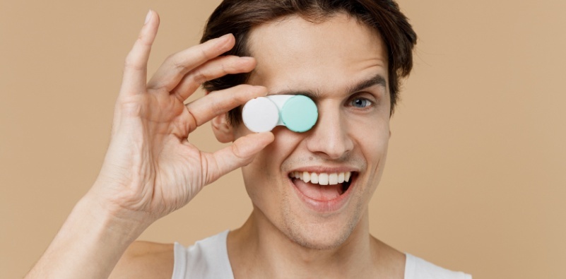A young man wearing a white vest holding a contact lens case covering his right eye.