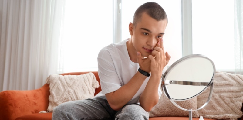 A young man sat on a sofa, looking in a mirror inserting a contact lens in his eye.