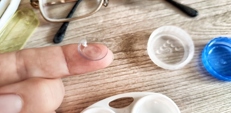 A person carefully holds a contact lens on their fingertip next to glasses and lens care products on a table.