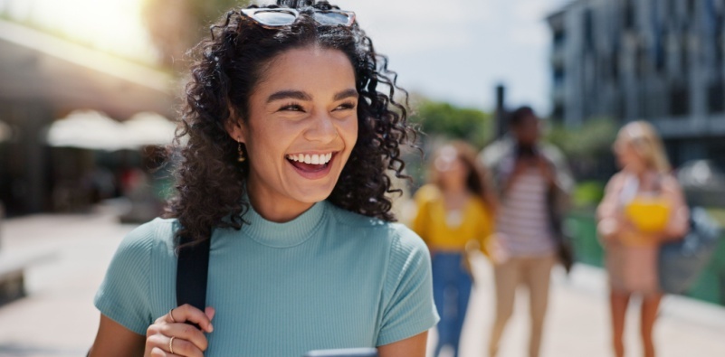 A smiling woman, outdoors, holding on to the strap of a backpack, with sunglasses on the top of her head.]