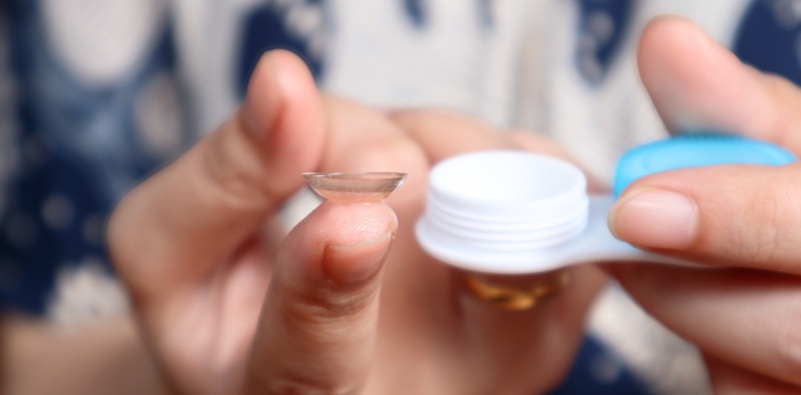 A contact lens on the fingertip of a young woman who is holding a contact lens storage container in her other hand.